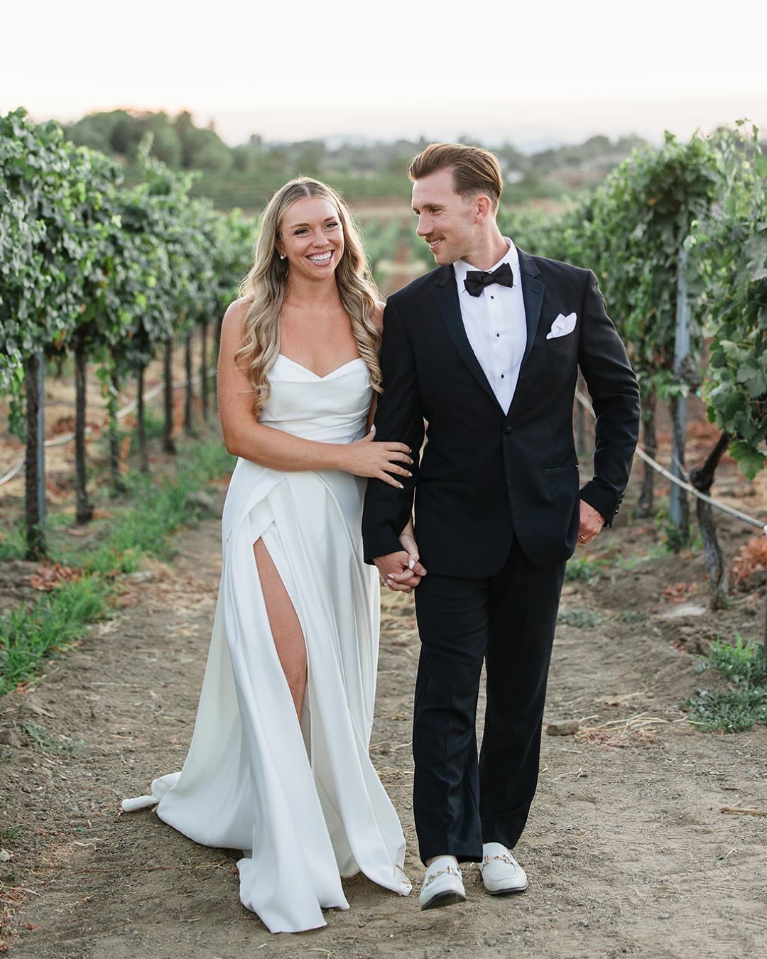 Smiling bride with long soft waves walking through a vineyard with her groom